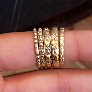 Puede incluir: Una colección de seis anillos dorados, cada uno con un diseño texturizado único. Los anillos varían en estilo, incluyendo motivos de cuentas, estampados y florales. Los anillos están apilados, mostrando sus diferentes texturas y detalles.