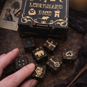 Peut inclure: Jeu de dés Lenormand en bois noir avec des détails dorés. Les dés présentent des symboles comme une clé, un ours et une croix. L'ensemble comprend une boîte avec "Lenormand Dice" et des motifs décoratifs. Une bougie allumée et une carte sont en arrière-plan.