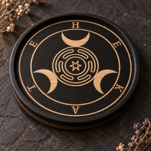 Puede incluir: Un posavasos circular de madera negra con un diseño grabado a láser. El centro muestra una estrella dentro de un patrón de laberinto, rodeada por tres lunas crecientes. Las letras H, E, K, A, T, E están espaciadas en el anillo exterior.
