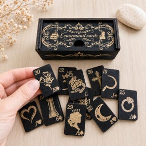 May include: A black wooden box with gold filigree and the text Lenormand cards, accompanied by a set of small black rectangular cards featuring gold illustrations of symbols like a heart, snake, ring, and crescent moon with numbers.