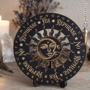 May include: Black and gold decorative planchette with a sun and moon design. The planchette has the words "Yes", "No", and "Rephrase" around the edge, along with the alphabet and numbers.
