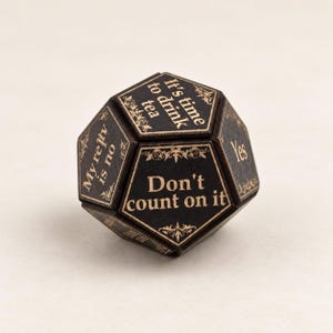 May include: A black dodecahedron-shaped decision die with gold-coloured engraved text and decorative filigree borders. Visible text includes 'It's time to drink tea', 'Don't count on it', 'Yes', and 'My reply is no' on the various pentagonal faces.
