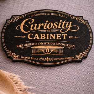 Può includere: Cartello nero e oro con la scritta "Curiosity Cabinet" in elegante corsivo. Il cartello include anche le frasi "Wonders & Oddities", "Rare Artifacts & Mysterious Discoveries" e "Strange Relics & Marvelous Specimens."