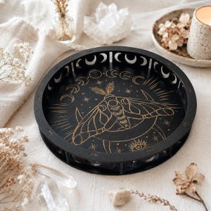 Peut inclure: Un plateau rond en bois noir orné d'une illustration de papillon de nuit dorée au centre, entourée de motifs célestes incluant des étoiles, un soleil rayonnant et un cycle de phases lunaires. Le rebord présente des découpes en forme de lune.