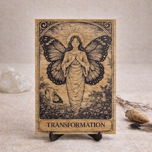 Peut inclure: Illustration détaillée sur bois d'une femme ailée avec des ailes de papillon, un halo et le mot "TRANSFORMATION". L'œuvre est présentée sur un petit support noir.