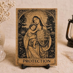 Puede incluir: Placa de madera con un grabado detallado de una madre y un niño, con un escudo con una cruz. La obra de arte está enmarcada por un borde adornado, con la palabra "PROTECTION" en la parte inferior. La placa se muestra en un soporte negro.