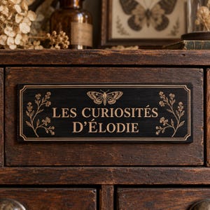 May include: A rectangular black wooden sign featuring the gold-colored text LES CURIOSITÉS D'ÉLODIE, centered with a butterfly illustration and flanked by botanical sprigs on both sides, all enclosed within a thin gold-colored border line.