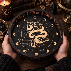 Könnte beinhalten: Ein rundes Tablett aus schwarzem Holz mit einer goldfarbenen Grafik einer gewundenen Schlange, die mit geometrischen Kristallformen verflochten ist. Der Rand des Tabletts zeigt eine kreisförmige Abfolge von Mondphasen und die Seitenwände ein Gittermuster.