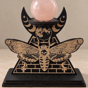 Pode incluir: Um suporte de exibição de madeira preta com uma mariposa com uma marca de caveira no corpo, uma lua cheia e fases da lua. O suporte tem uma plataforma circular no topo que sustenta uma esfera de cristal lisa de cor rosa pálido.
