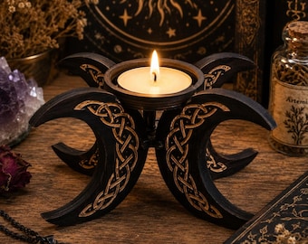 May include: A black wooden candle holder featuring a triple moon design with intricate gold-colored Celtic knotwork patterns. The central circular cup holds a single lit white tea light candle, creating a warm glow against the dark wood finish.