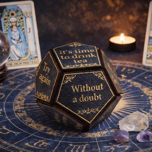 Könnte beinhalten: Eine dunkelblaue, vielseitige dekorative Box mit goldfarbenen Schriftzügen, die lauten: „Es ist Zeit, Tee zu trinken“ und „Ohne Zweifel“. Die Box ist von Tarotkarten, Kristallen und einer brennenden Kerze umgeben. Der Hintergrund ist eine blaue astrologische Karte.