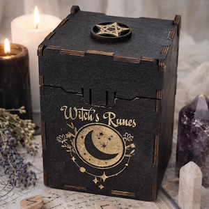 Puede incluir: Caja de madera negra con el texto "Witch's Runes" y un diseño de media luna. La caja está adornada con un emblema en forma de estrella en la tapa. Se ven velas, cristales y runas de madera.