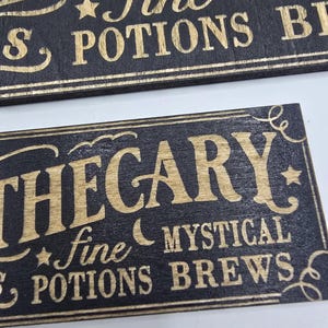 May include: Two rectangular wooden signs with gold lettering on a black background. The top sign reads "fine POTIONS BREWS" and the bottom sign reads "APOTHECARY fine MYSTICAL POTIONS BREWS". Decorative stars and crescent moons are included.