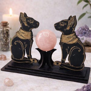 May include: A black wooden ornament with two cat figures in a seated pose, adorned with gold detailing, either side of a pink crystal ball. The cats sit on a black base, with a candle and crystals in the background.
