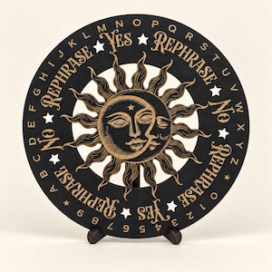 May include: A circular black board featuring a central sun and moon illustration with gold-toned details. The outer edge displays the alphabet from A to Z, numbers 0 to 9, and the words YES, NO, and REPHRASE printed in a decorative font.