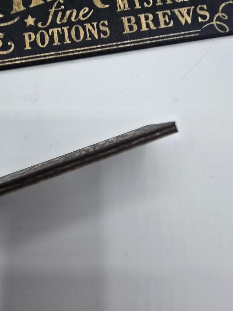 May include: A black wooden sign with gold lettering that reads "fine potions brews". The sign has a dark, textured edge. The background is white.