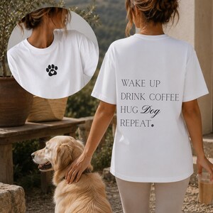 Könnte beinhalten: Ein weißes Kurzarm-T-Shirt mit einem kleinen schwarzen Pfotenabdruck mit einem Herz in der Mitte auf der Vorderseite. Die Rückseite zeigt den schwarzen Text: WAKE UP, DRINK COFFEE, HUG Dog, REPEAT, mit einem kleinen Pfoten-Symbol am Zeilenende.