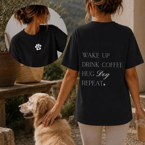 Könnte beinhalten: Ein schwarzes Kurzarm-T-Shirt mit einem kleinen weißen Pfotenabdruck auf der Brust. Auf der Rückseite des Shirts steht der weiße Text: WAKE UP, DRINK COFFEE, HUG Dog, REPEAT, ergänzt durch ein kleines Pfotenabdruck-Symbol am Ende der Zeile.