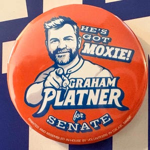 May include: A round, orange campaign button with a portrait of a man pointing. Text on the button reads "HE'S GOT MOXIE!" and "GRAHAM PLATNER for SENATE". The button is printed in white and blue.