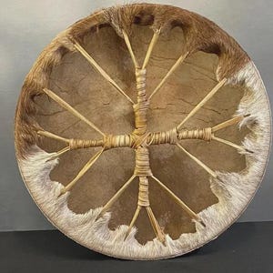 Op de afbeelding: Een ronde frametrommel met een strak gespannen, lichtbruin dierenhuid oppervlak. De trommel is afgewerkt met een dikke, harige rand in bruin en wit. Een centraal kruisvormig vetersysteem van lichte stroken houdt de huid op zijn plek.