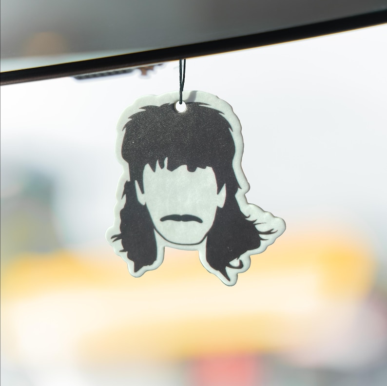 Puede incluir: Ambientador en blanco y negro con forma de corte de pelo mullet. Cuelga de un cord&oacute;n negro. El fondo est&aacute; borroso, con suaves tonos amarillos y blancos.