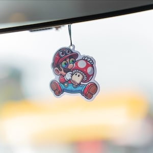 May include: A colorful air freshener featuring a cartoon character in a red hat and overalls, holding a red and white mushroom. The character has rainbow-colored eyes and is sitting down. The air freshener is hanging from a black string.