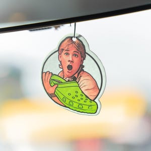 May include: A car air freshener with a cartoon image of a person holding a green Croc shoe, looking surprised. The air freshener is round with a white border and a black string for hanging.