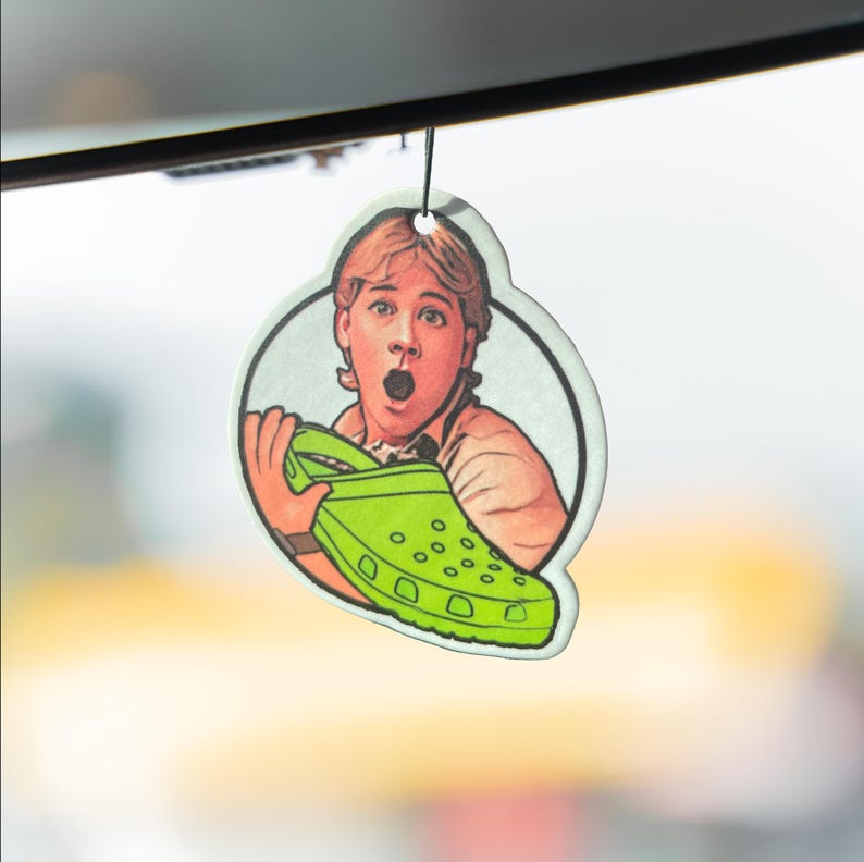 May include: A car air freshener with a cartoon image of a person holding a green Croc shoe, looking surprised. The air freshener is round with a white border and a black string for hanging.
