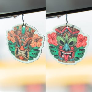 Op de afbeelding: Twee auto-luchtverfrissers met een tiki-maskerontwerp. De maskers zijn in tinten bruin, oranje, groen en blauw, met rode hibiscusbloemen en groene bladeren. Elke luchtverfrisser heeft een zwart touwtje om op te hangen.