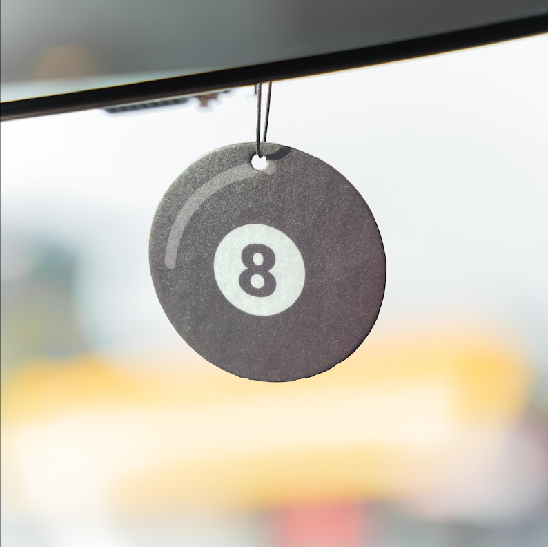 May include: A round, dark gray air freshener with a white number 8, resembling a billiard ball. The air freshener is hanging by a black string. The background is blurred, suggesting it's hanging in a vehicle.