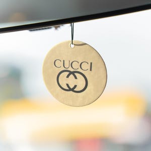 May include: A round, tan tag with the word "CUCCI" printed in black, above a black interlocking double-C logo. The tag is suspended by a thin black string. The background is blurred, with a hint of a window frame.