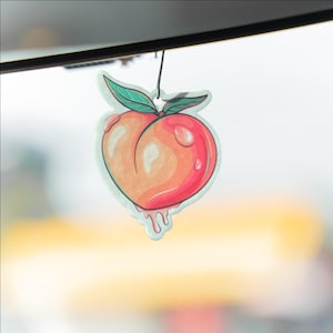 May include: A peach-shaped air freshener with a watercolor design. The peach is a mix of orange and red, with green leaves and a black string for hanging. The design includes water droplets and dripping juice.
