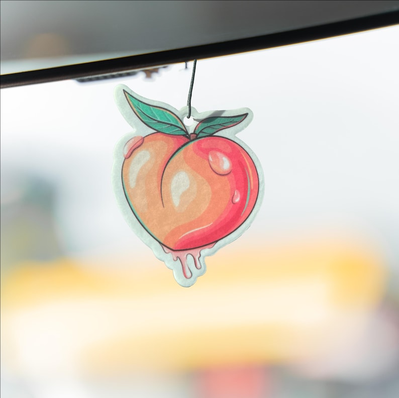 May include: A peach-shaped air freshener with a watercolor design. The peach is a mix of orange and red, with green leaves and a black string for hanging. The design includes water droplets and dripping juice.