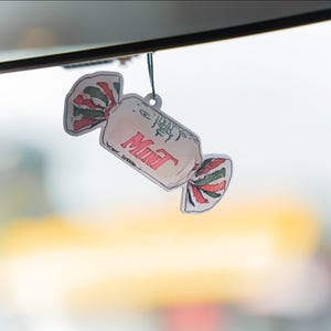 May include: A candy-shaped air freshener with the word "Mint" in red. The wrapper design features red, white, and green accents. The air freshener is hanging from a black string.