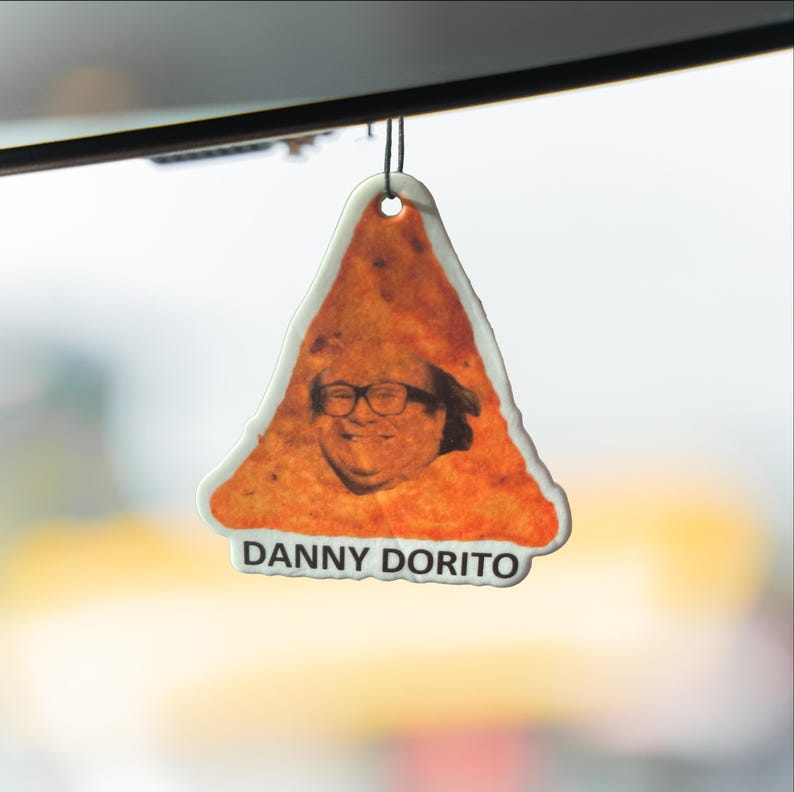 Puede incluir: Un ambientador triangular con un retrato de Danny DeVito como una patata frita Dorito. El ambientador tiene un borde blanco y el texto "DANNY DORITO" en la parte inferior. El fondo est&aacute; borroso.