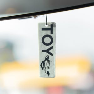 May include: A rectangular, off-white tag with the word "TOY" printed vertically in black. Below the text is a black illustration of a horse's head. The tag is suspended by a black string.