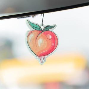 May include: A peach-shaped air freshener hanging from a black string. The design features a gradient of orange and red with two green leaves at the top. The bottom of the peach has a light blue dripping effect, and the surface includes white highlights.