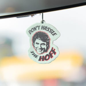 Puede incluir: Un ambientador verde claro con una cuerda negra. Presenta una cara sonriente y el texto "DON'T HASSLE THE HOFF" en rojo y negro.
