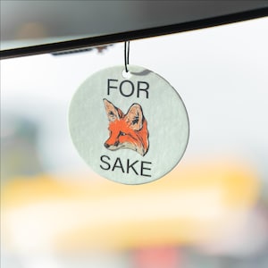 May include: A round, light green air freshener with the words "FOR SAKE" printed in black. The air freshener features an orange and white fox illustration. It is hanging by a black string.