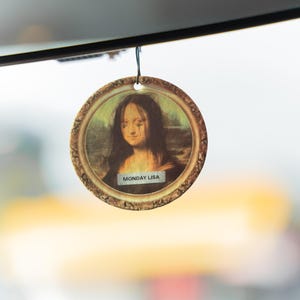 May include: Round air freshener featuring a parody of the Mona Lisa painting with tears, set in a gold frame. The text "MONDAY LISA" is printed at the bottom. The air freshener is hanging from a black string.