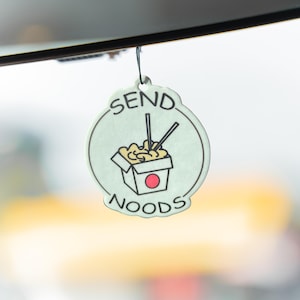 Op de afbeelding: Een lichtgroene luchtverfrisser met een zwart touwtje, met een cartoonillustratie van een noedelbox met eetstokjes. De tekst "SEND NOODS" is rondom de afbeelding geschreven in een cirkelvormig ontwerp.