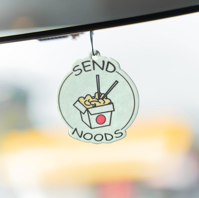 Op de afbeelding: Een lichtgroene luchtverfrisser met een zwart touwtje, met een cartoonillustratie van een noedelbox met eetstokjes. De tekst "SEND NOODS" is rondom de afbeelding geschreven in een cirkelvormig ontwerp.