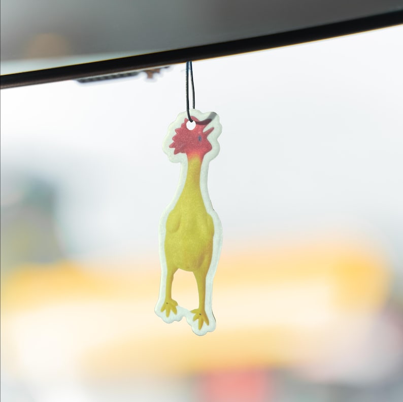 May include: A yellow and red chicken-shaped air freshener hanging from a black string. The air freshener has a cartoonish design with a yellow body, red comb, and a white border. The background is blurred.