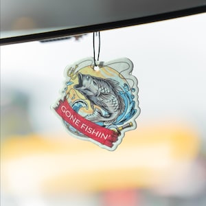 以下が含まれることがあります： 赤いバナーに「GONE FISHIN'」の文字と魚のグラフィックが描かれた芳香剤。デザインは、跳ねる魚、水、釣り道具が特徴です。芳香剤は黒い紐で吊り下げられています。