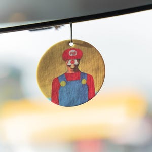 May include: A round, yellow air freshener featuring a cartoon illustration of a character in a red hat, red shirt, and blue overalls. The character's face is partially obscured. The air freshener is hanging from a black string.