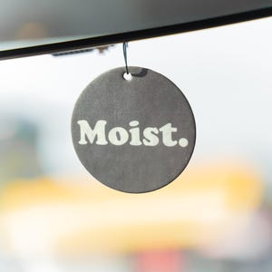 May include: A circular, grey air freshener with the word "Moist." in white lettering. It hangs from a black string. The air freshener has a small hole at the top. The background is blurred, with a hint of yellow.
