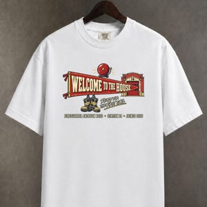May include: A white short-sleeve t-shirt featuring a graphic of a red pennant with the text "WELCOME TO THE HOUSE," a red fire alarm bell, a fire station building labeled "14," and black firefighter boots with the text "READY FOR THE FIRST CALL."