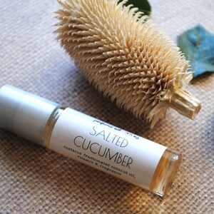 May include: A clear glass roll-on bottle with a silver cap, labeled "Salted Cucumber." The bottle is next to a dried, textured plant and two green leaves. The label lists ingredients.