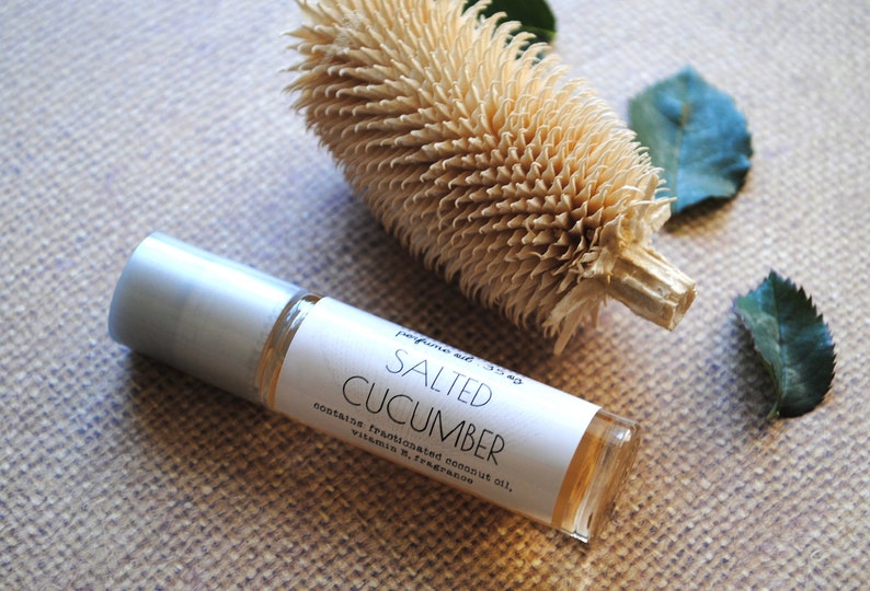 May include: A clear glass roll-on bottle with a silver cap, labeled "Salted Cucumber." The bottle is next to a dried, textured plant and two green leaves. The label lists ingredients.