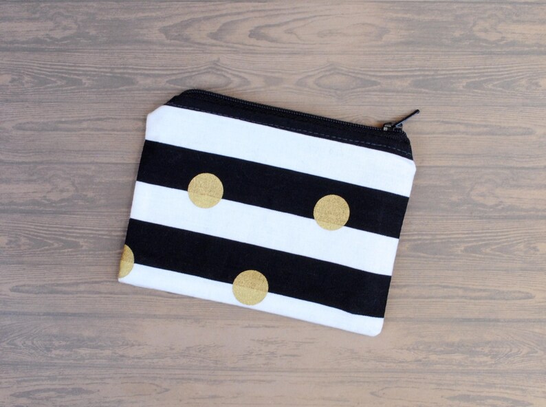 May include: A small, rectangular zippered pouch with a black and white striped pattern. The pouch features three gold-colored polka dots and a black zipper. The background is a light brown wooden surface.